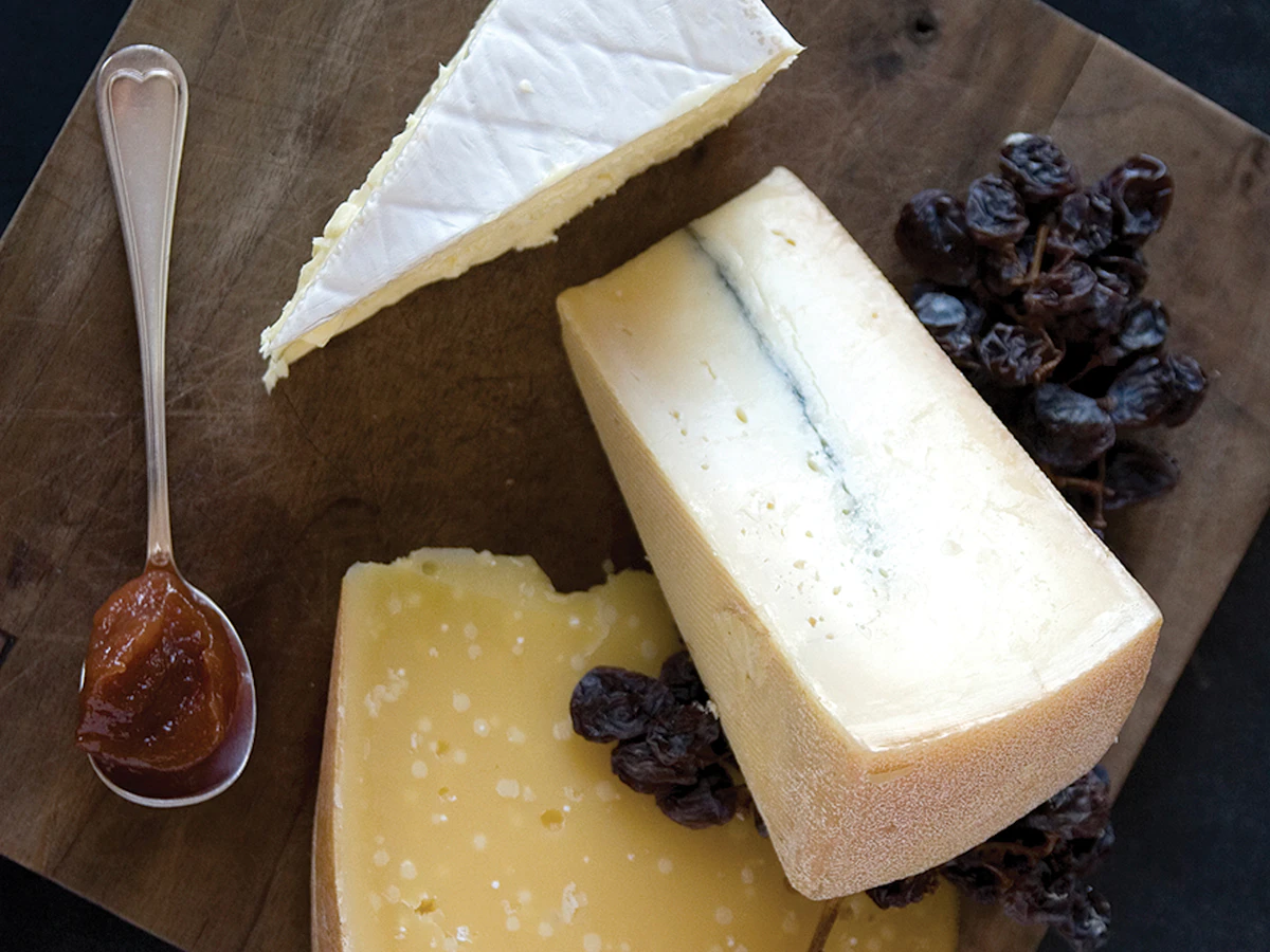 Three wedges of cheese on a dark wooden board with dried grapes and a spoonful of fruit preserve.