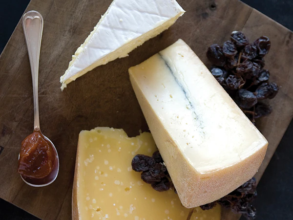 Three wedges of cheese on a dark wooden board with dried grapes and a spoonful of fruit preserve.