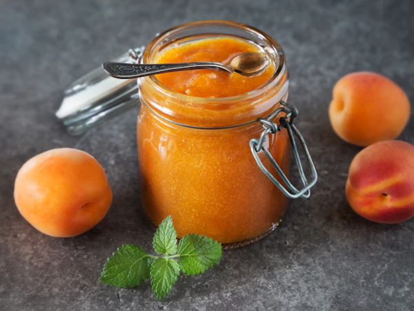 An open jar of apricot jam with a spoon, beside fresh apricots.