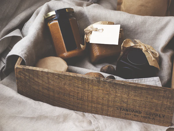 A wooden crate lined with linen, holding jars of preserves tied with twine and a blank gift tag.